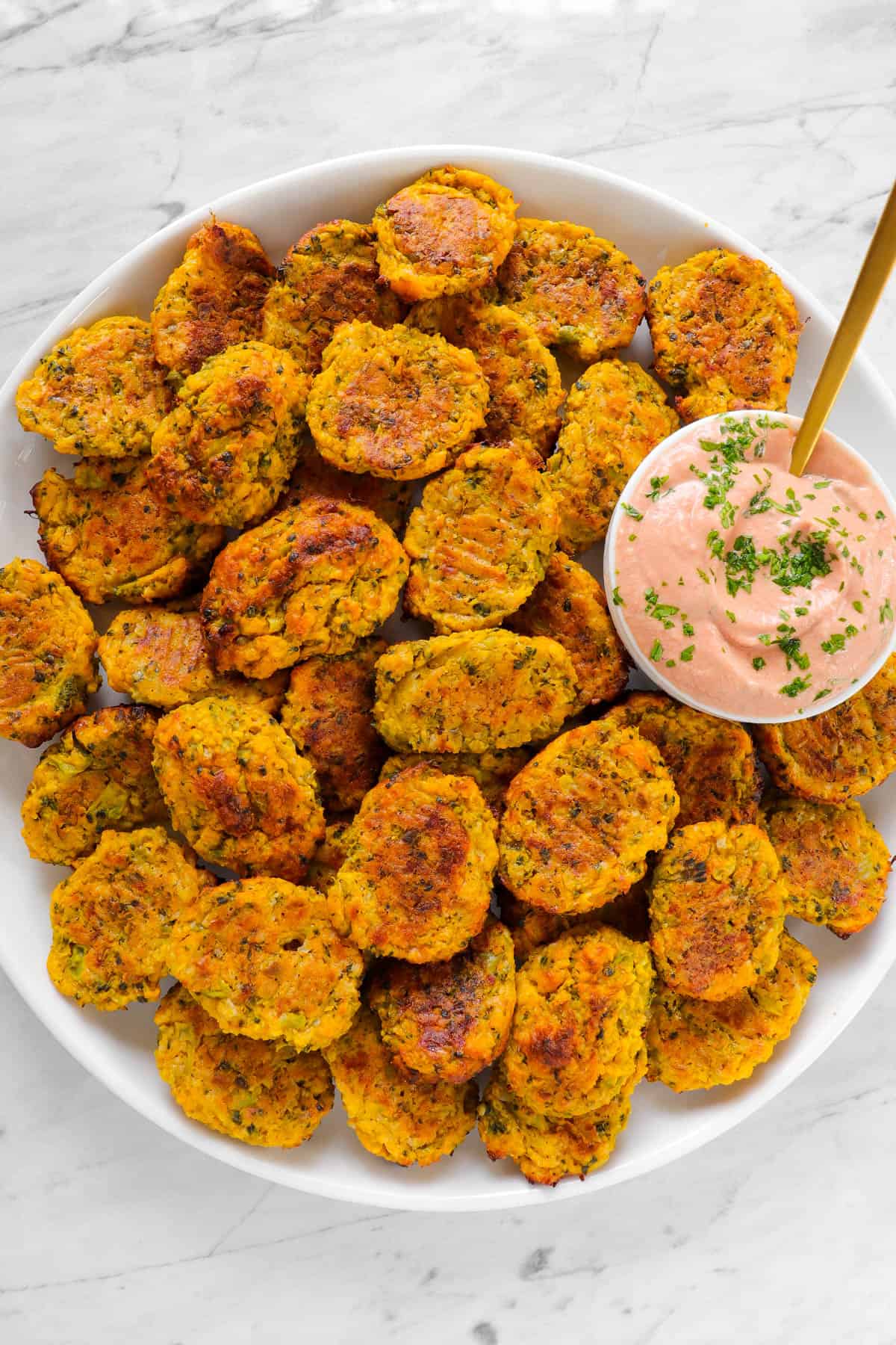 Baked broccoli pumpkin tots on a plate with tomato yoghurt sauce and golden spoon.