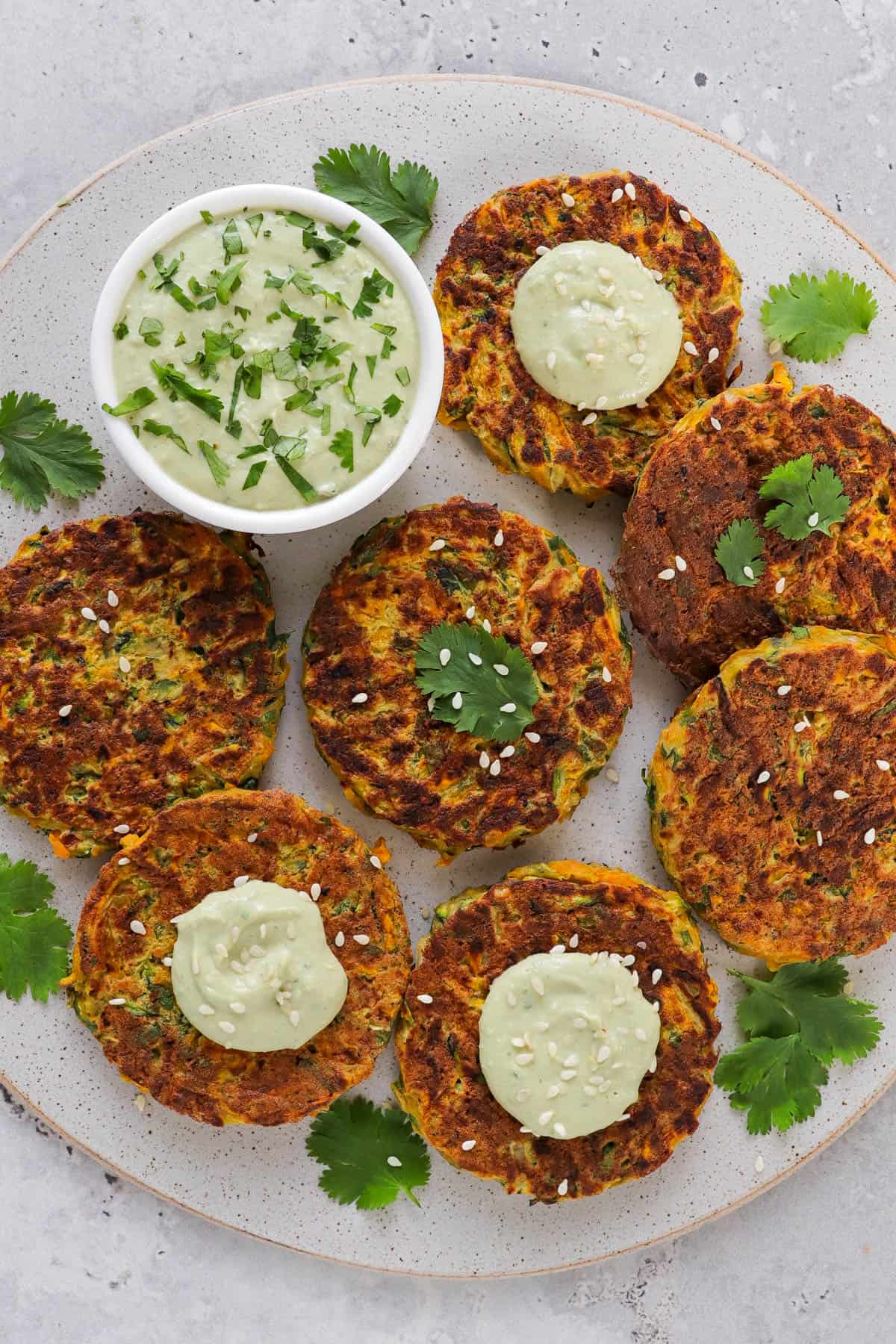 Cooked fritters on a plate with coriander leaves, sesame seeds and avocado dip on the side.