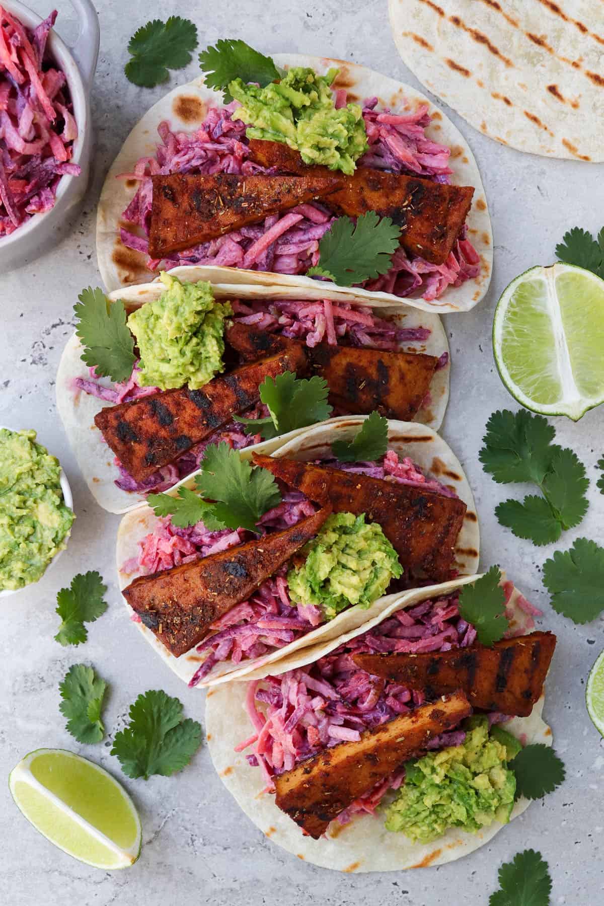 Top view of tofu tacos. Coriander leaves, limes, avocado and slaw on the sides.