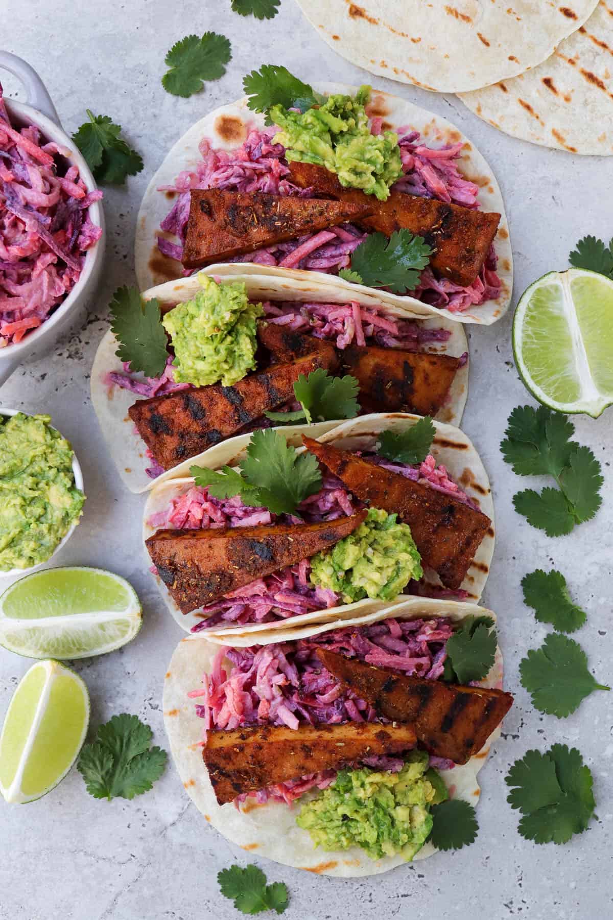 Top view of blackened tofu tacos. Coriander leaves, limes, avocado and slaw on the sides.