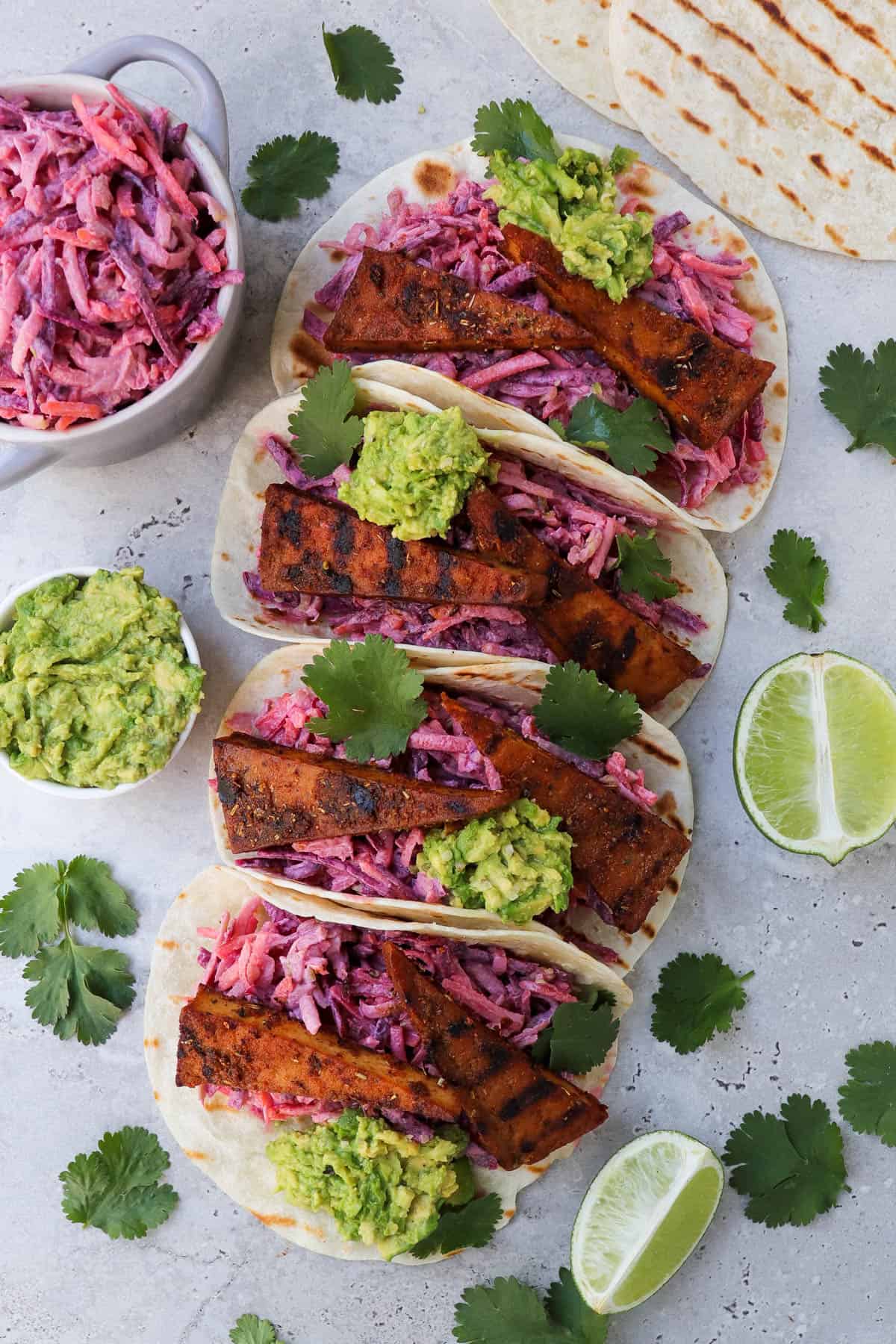 Top view of tofu tacos on angle. Coriander leaves, limes, avocado and slaw in a dish on the sides.