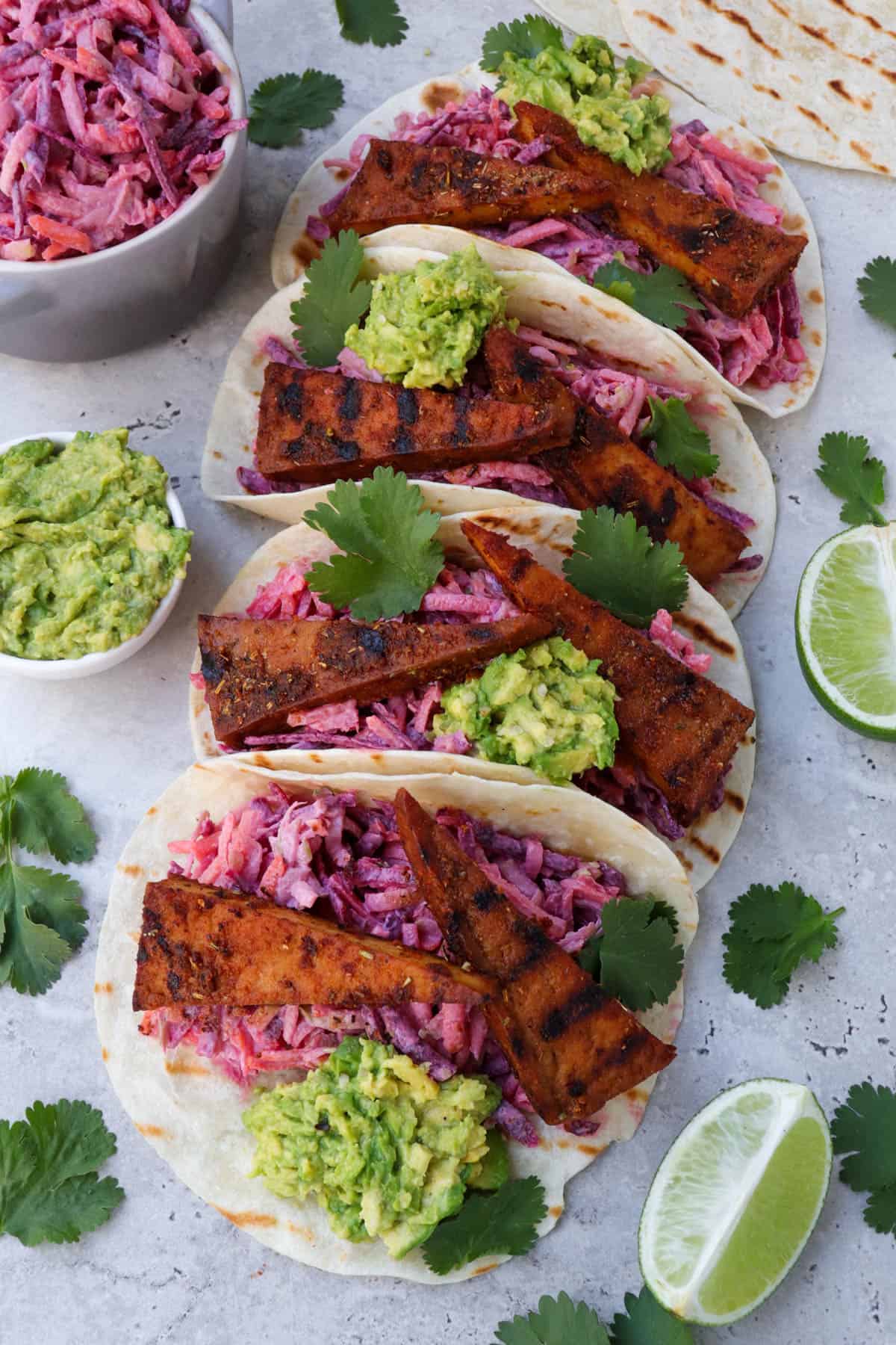 Blackened tofu tacos on angle. Coriander leaves, limes, avocado and slaw in a dish on the sides.