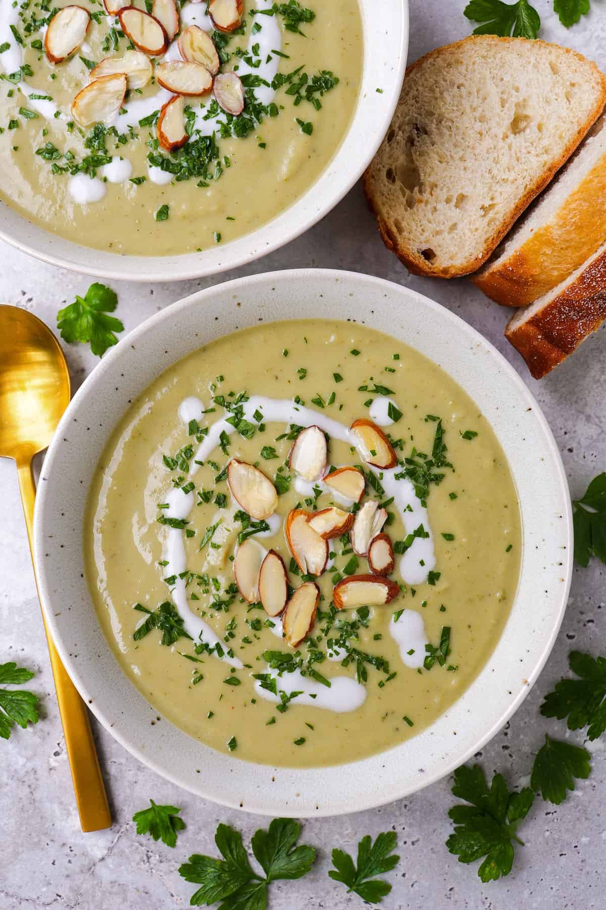 Broccoli soup in a bowl topped with coconut cream. almond flakes, finely chopped parsley. Sliced up sourdough o the side.