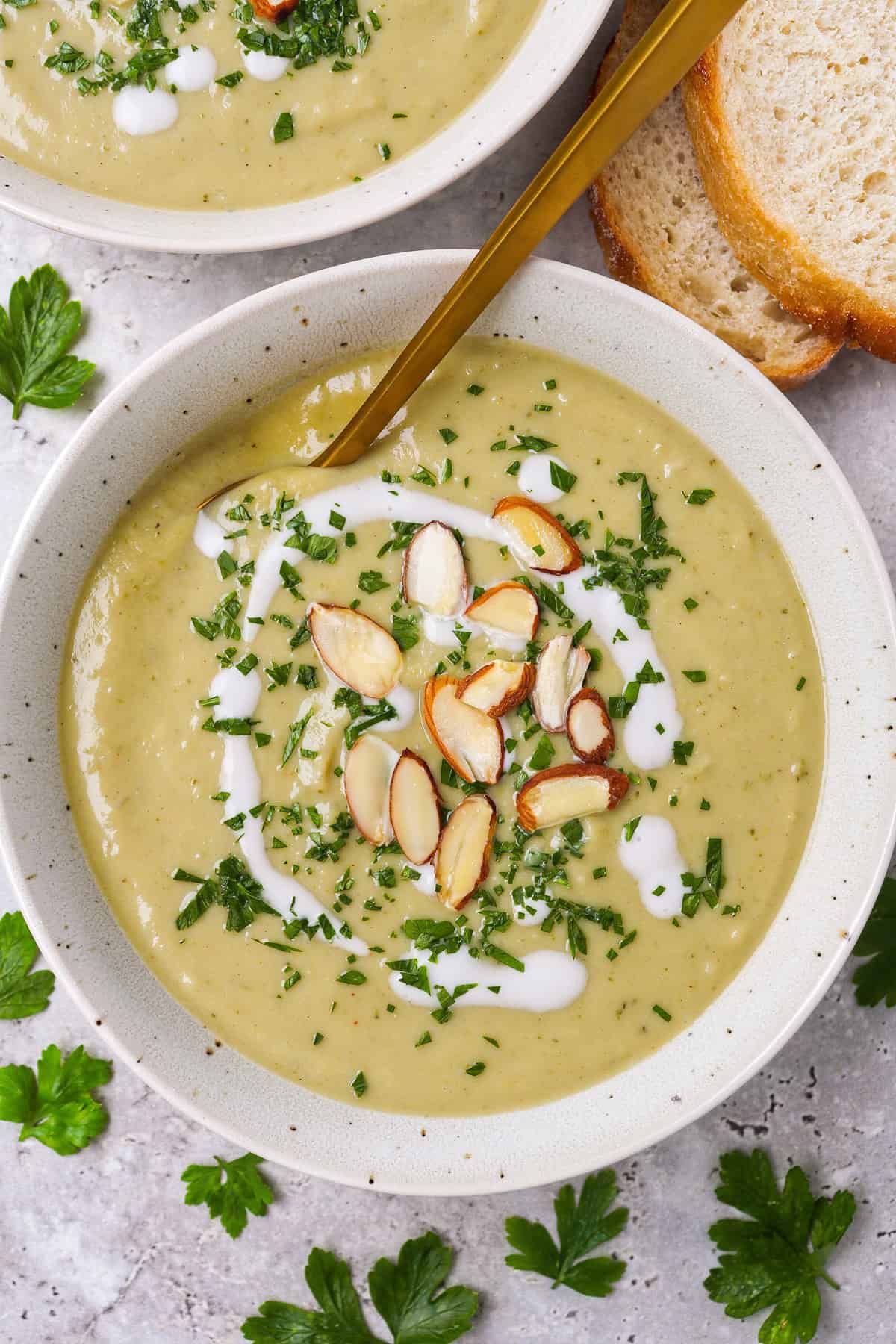 Broccoli soup in a bowl with a gold spoon dipped in. Topped with coconut cream. almond flakes, finely chopped parsley.