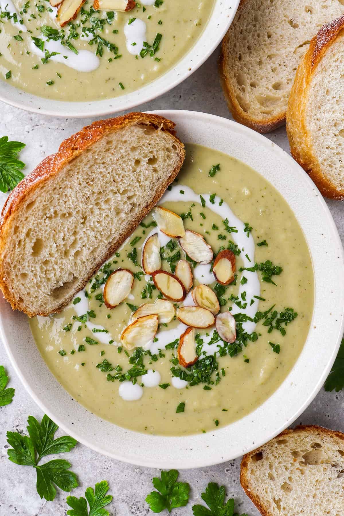 Broccoli soup in a bowl topped with coconut cream. almond flakes, finely chopped parsley and a slice of sourdough bread.