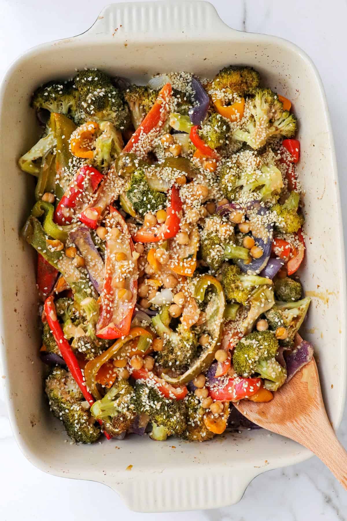 Baked chickpeas and veggies in dish with sesame seeds and wooden spoon.