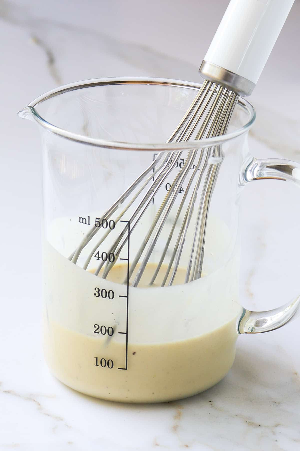 Tahini dressing in a glass jug with a white whisk.
