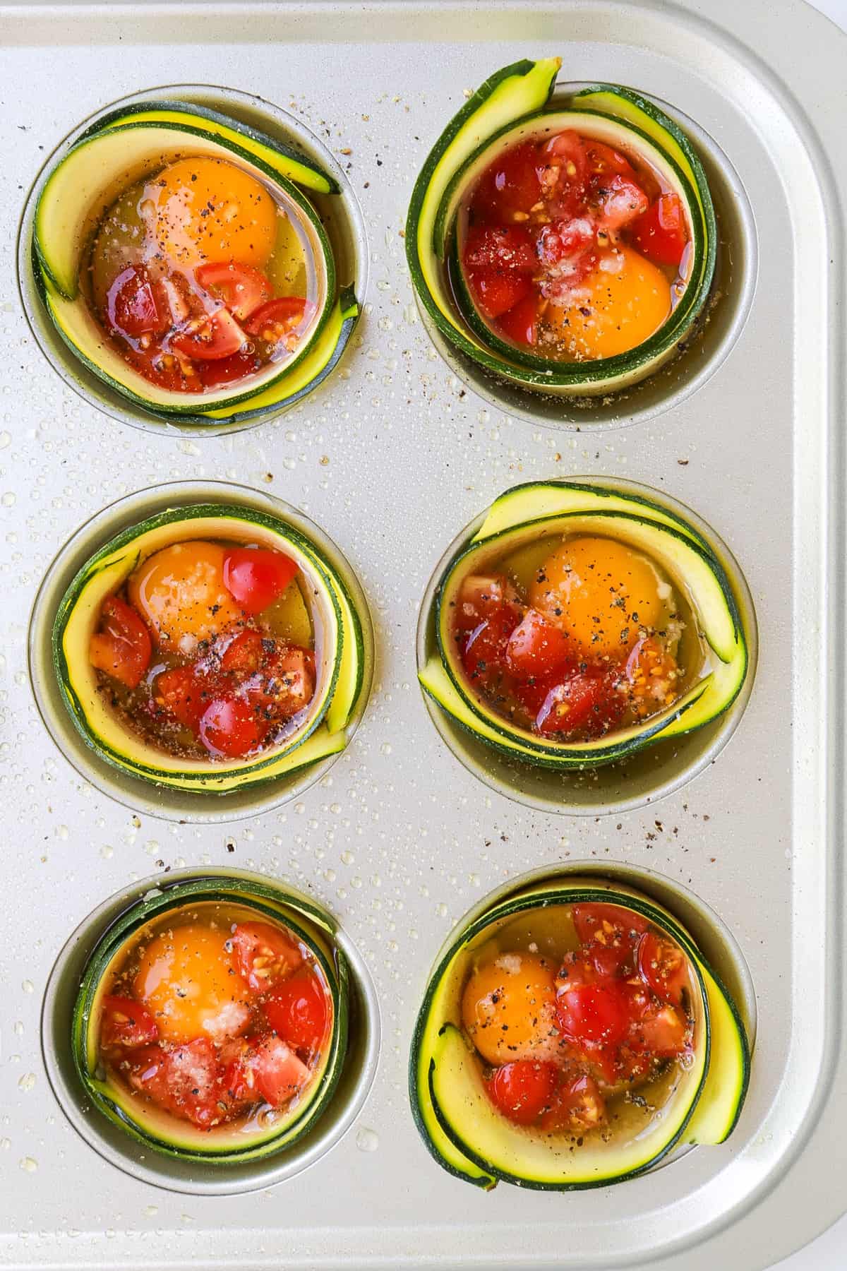 Muffin pan with raw zucchini strips wrapped around each cavity filled with egg and tomatoes.
