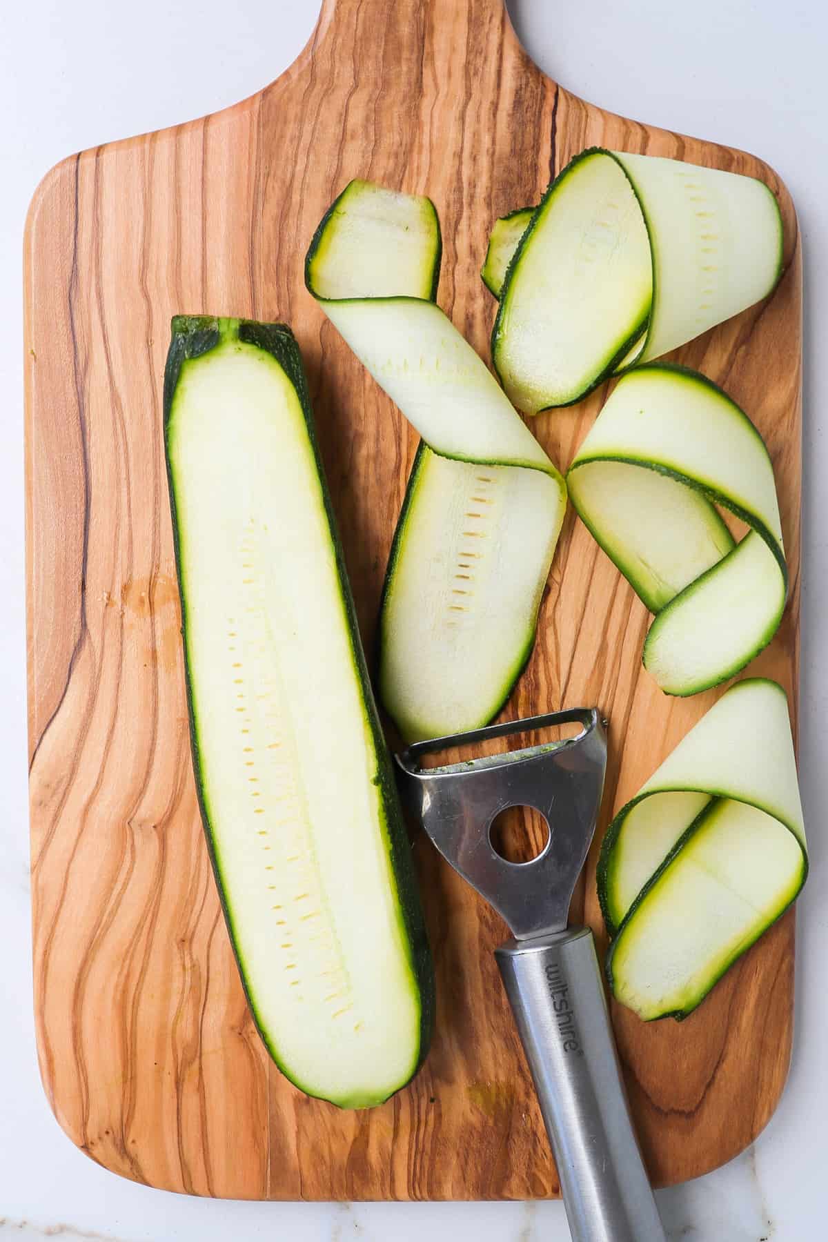 A zucchini and strips of zucchini on a wooden cutting board with a vegetable peeler.
