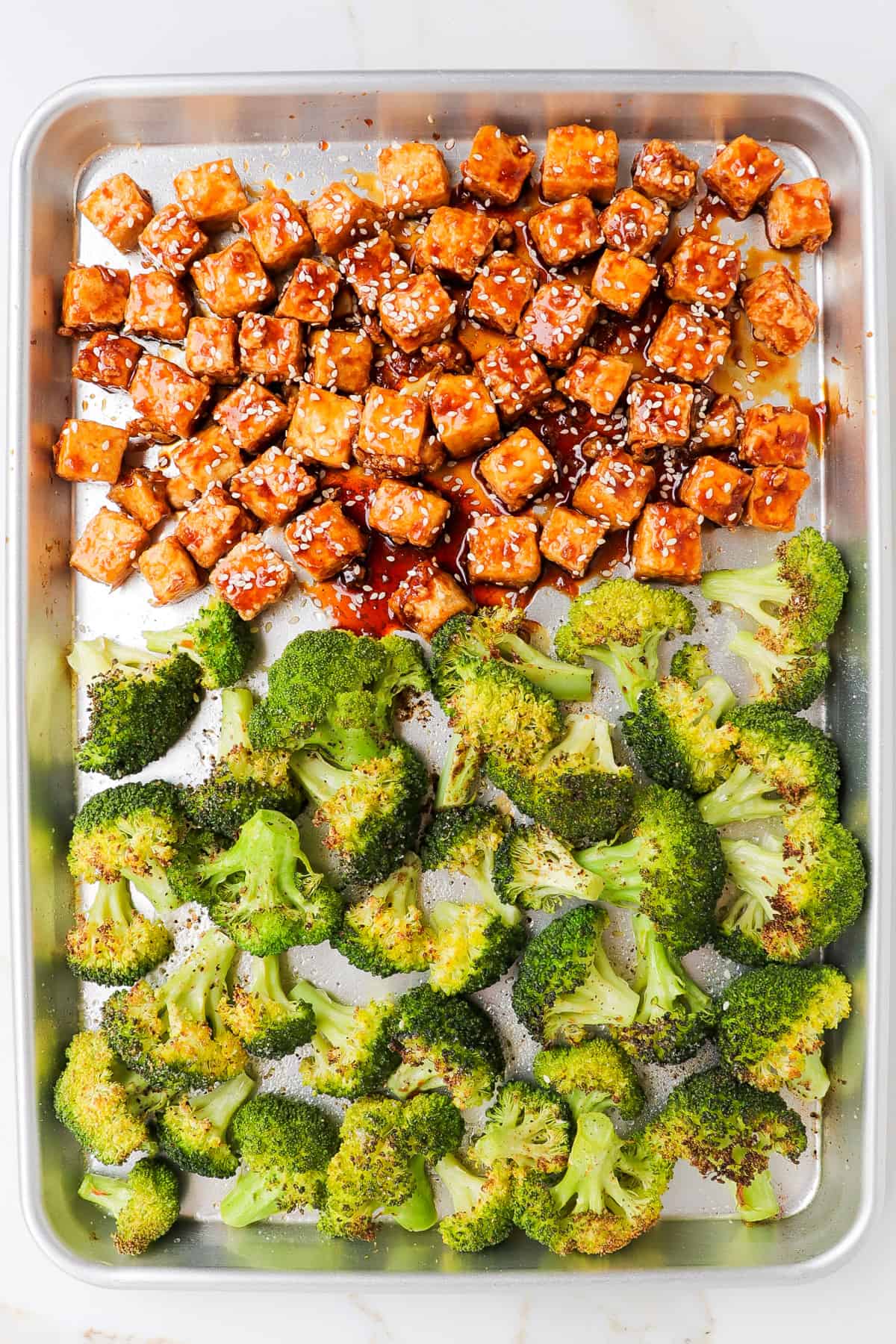 Tofu coated in honey garlic sauce with sesame seeds and broccoli on a pan.