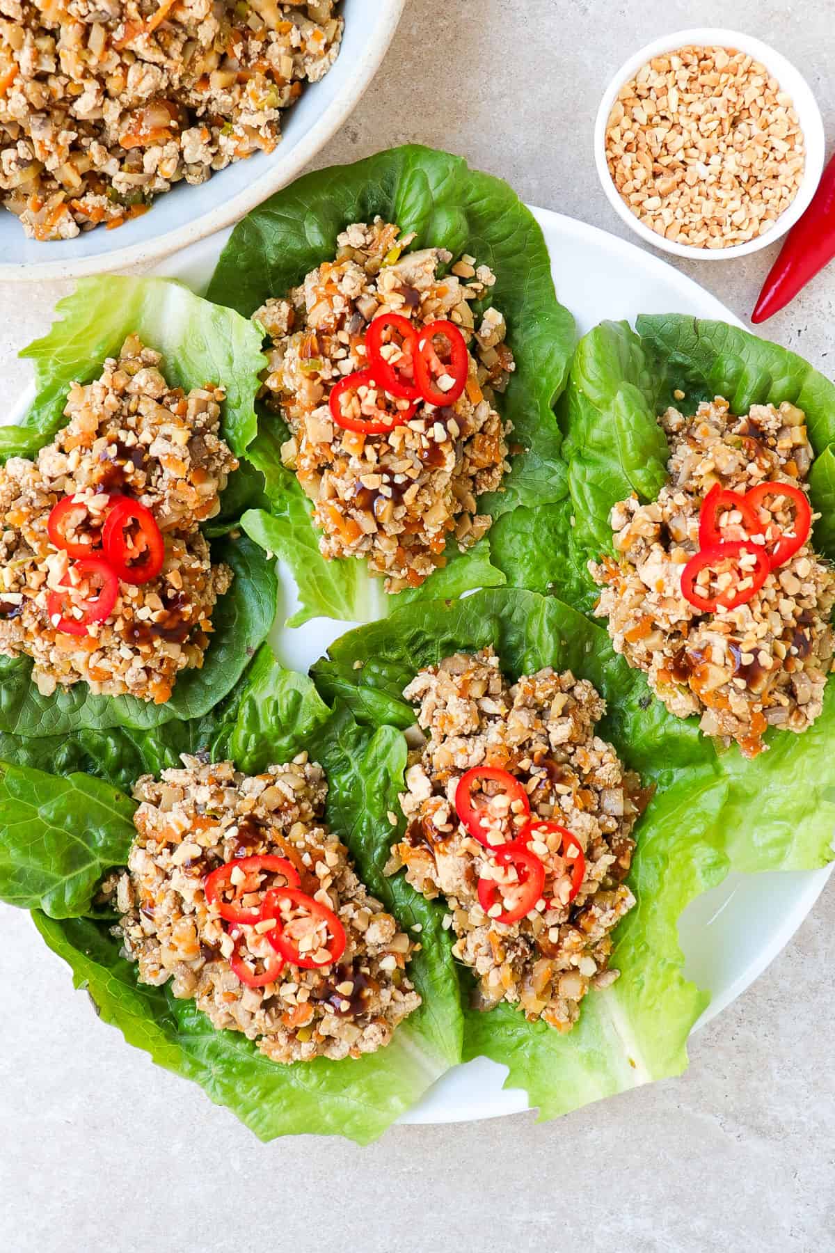 Five lettuce wraps on a plate topped with sliced chilli and crushed peanuts. Extra tofu filling is inn a bowl on the side.