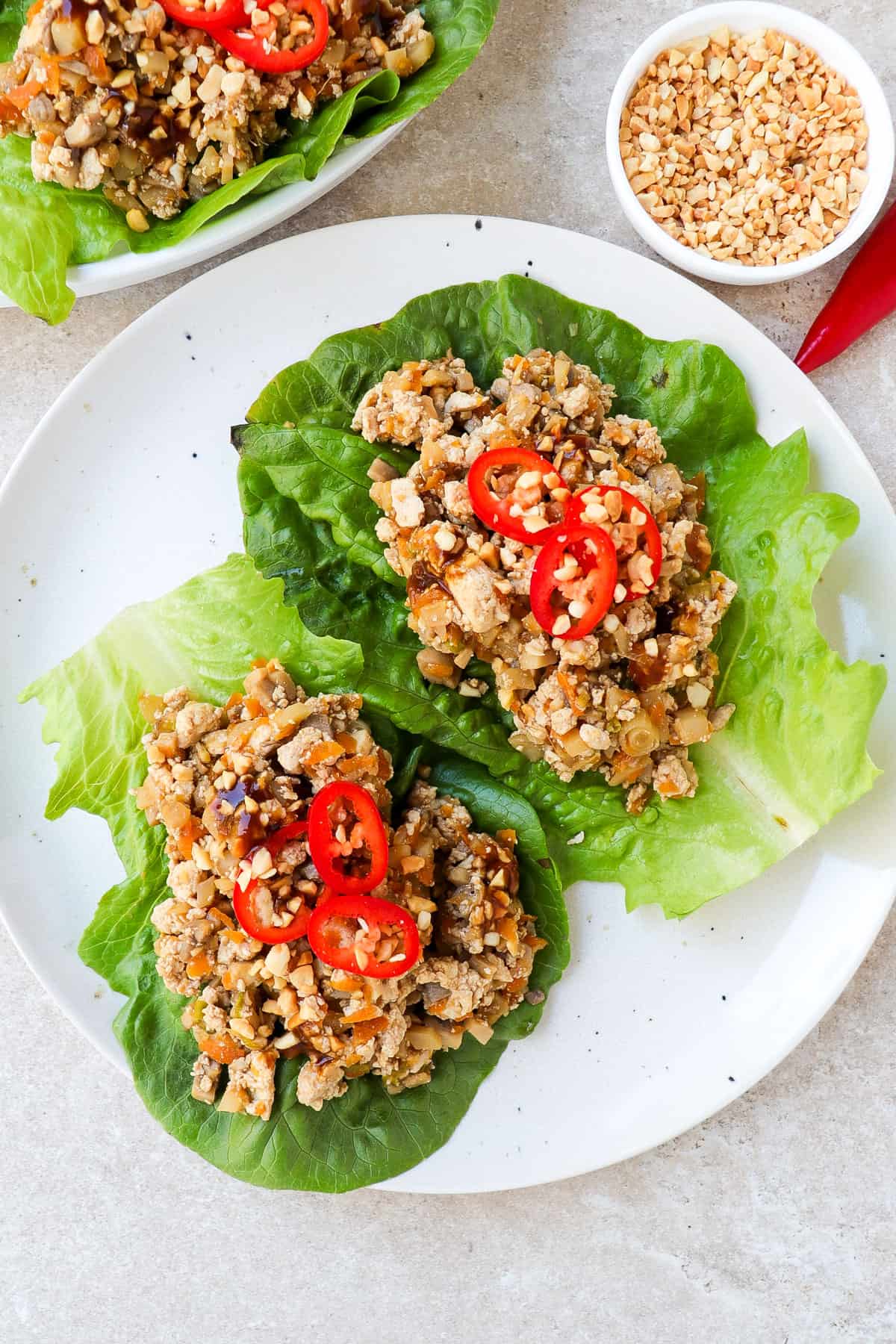 Vegan san choy bow lettuce wraps on a plate topped with sliced chilli and crushed peanuts. Extra tofu filling is inn a bowl on the side.