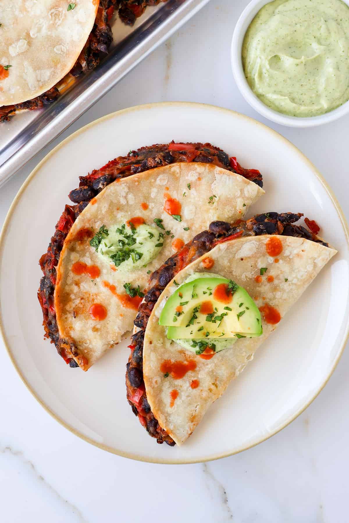 Two tacos on a plate topped with avocado, crema and hot sauce.